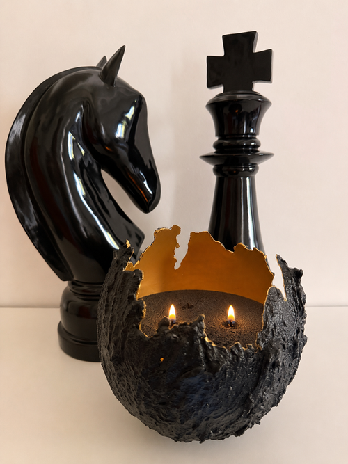 Bougie sablée Noir Profond allumée dans un contenant noir texturé avec intérieur doré, mise en scène élégante avec pièces d’échecs noires.