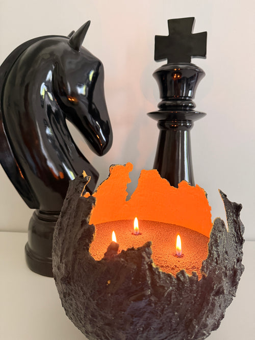 Bougie sablée Noir Profond Maison Charlee allumée dans un œuf de dragon en béton noir, intérieur orange, avec pièces d’échecs noires